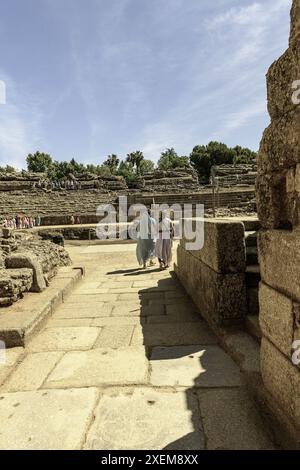 Due donne vestite in abiti romani camminano attraverso uno storico arco in pietra a Merida, in Spagna, con l'antico anfiteatro visibile sullo sfondo. Foto Stock