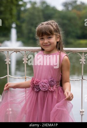 Una ragazza dolce e sorridente di 6 anni con capelli lunghi e scuri guarda allegramente la fotocamera. Vestito con un bellissimo vestito rosa. Tiene i bordi del vestito. Foto Stock