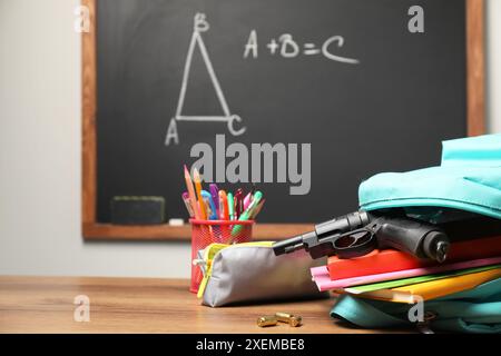 Pistola, proiettili e cancelleria scolastica su un tavolo di legno vicino alla lavagna interna, spazio per il testo Foto Stock