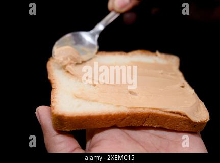 Una foto ravvicinata di una deliziosa crema di burro di arachidi che viene versato su una fetta di pane tostato con un cucchiaio di metallo Foto Stock