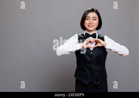 Servizio catering donna dipendente vestita con uniforme da cameriera che mostra il simbolo del cuore con il ritratto delle dita. Lavoratore del ristorante che dimostra il gesto d'amore e guarda la fotocamera Foto Stock