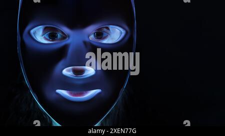 Uno scatto ravvicinato di una donna che indossa una maschera fototerapica su sfondo nero. La maschera le sta illuminando il viso e i suoi occhi sono visibili dietro la maschera. Foto Stock