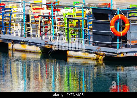 una fila di sedie colorate si trova su un molo accanto a un salvagente. Le sedie sono disposte in modo che siano rivolte verso l'acqua. Tranquillo bacino con Foto Stock