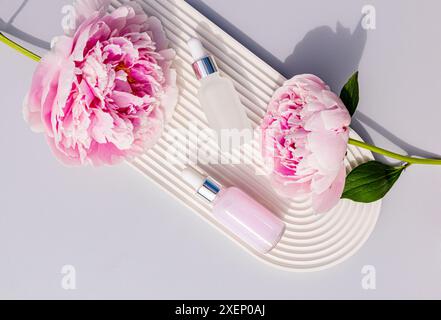 Due flaconi cosmetici opachi con contagocce e siero per la cura della pelle del viso e del corpo su un vassoio ad arco bianco goffrato e peonie. vista dall'alto. Foto Stock