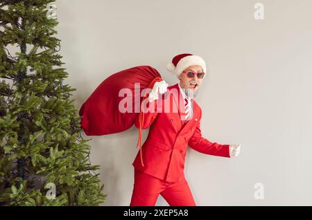 Un uomo allegro ed elegante con cappello di Babbo Natale porta una borsa rossa con regali di Natale sulle spalle. Foto Stock