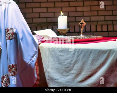 sacerdote ortodosso nell'altare che legge dal crocifisso biblico e accende la candela sul tavolo Foto Stock