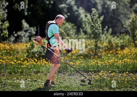 La sospensione dello zaino per il rifinitore per erbacce a benzina è indossata da un uomo alto che sta tagliando i leoni sul suo terreno. Foto Stock