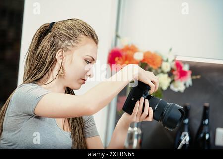 Una giovane donna con lunghe trecce sta regolando le impostazioni della sua fotocamera reflex digitale, preparandosi per una sessione di fotografia gastronomica in una cucina moderna Foto Stock