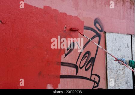 Processo di un pittore in tute verdi mentre dipinge su graffiti su una parete rossa, coprendo vecchie etichette con vernice fresca da un rullo Foto Stock