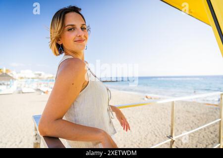 Una donna è in piedi su una spiaggia con un ombrello giallo dietro di lei. Sta sorridendo e posa per un film Foto Stock