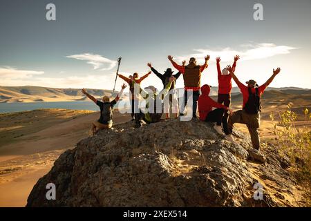 Un grande e variegato gruppo di escursionisti o turisti felici si trova sulla cima della montagna a braccia aperte contro il deserto del tramonto e il grande lago Foto Stock