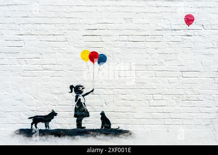 Banksy ispirò un dipinto in bianco e nero su una parete di una bambina con cane e gatto, tenendo in mano tre palloncini colorati e un quarto galleggiante libero. Foto Stock