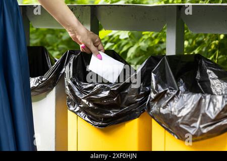la mano di una donna getta un foglio di carta piegato nella spazzatura. donna che getta via fogli di carta piegati. cestino nel parco. Foto di alta qualità Foto Stock