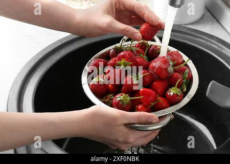 Donna che lava fragole fresche sotto l'acqua corrente in un colino metallico sopra il lavandino, primo piano Foto Stock