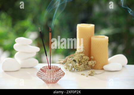 Bastoncini di incenso che si mescolano nel supporto vicino a pietre, candele e fiori secchi su un tavolo di legno all'aperto Foto Stock