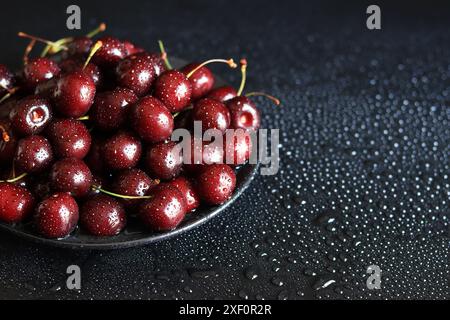 Ciliegie in un piatto nero su sfondo scuro e bagnato, primo piano. Ciliegie mature dolci bagnate. Ciliegie rosse scure con gambi verdi ricoperte da schizzi sottili o Foto Stock