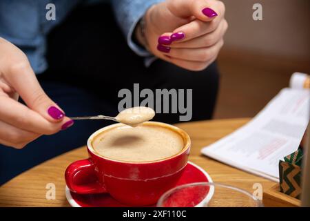 Primo piano di una mano di una donna che tiene un cucchiaio piccolo con schiuma di schiuma sopra l'espresso in una tazza rossa su un tavolo da caffè Foto Stock