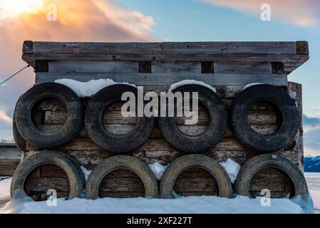 Grandi blocchi di ghiaccio ghiacciati su un porticciolo con pneumatici per auto in inverno. Il tramonto dorato si illumina sul lato di un lago gelido in inverno con il tramonto sullo sfondo. Foto Stock