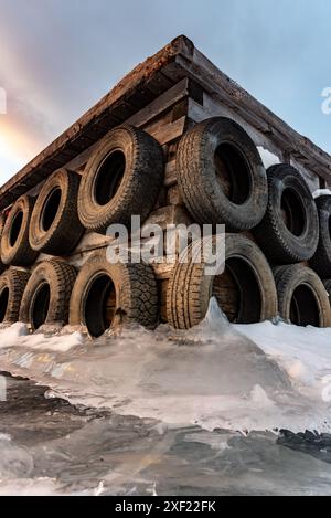 Grandi blocchi di ghiaccio ghiacciati su un porticciolo con pneumatici per auto in inverno. Il tramonto dorato si illumina sul lato di un lago gelido in inverno con il tramonto sullo sfondo. Foto Stock