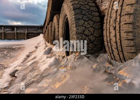 Grandi blocchi di ghiaccio ghiacciati su un porticciolo con pneumatici per auto in inverno. Il tramonto dorato si illumina sul lato di un lago gelido in inverno con il tramonto sullo sfondo. Foto Stock