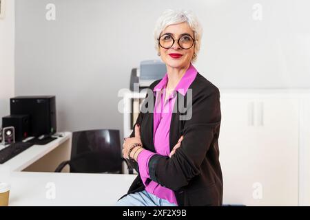 Una donna d'affari anziana con capelli e occhiali bianchi corti, che posa con sicurezza in un ambiente d'ufficio. Dirigente femminile professionale e di successo. Foto Stock