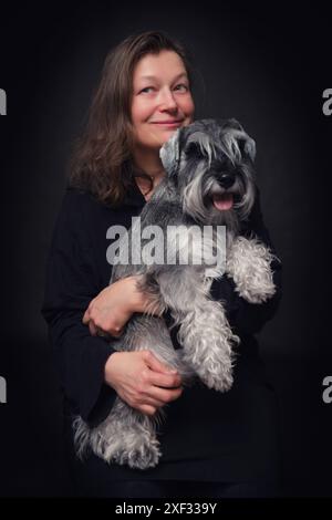 Una donna tiene in braccio un piccolo cane zwergschnauzer. Il cane è uno zwergschnauzer ed è felice. La donna sorride e sembra che si stia divertendo Foto Stock