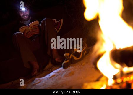 Un uomo legge il suo libro con un faro mentre è seduto intorno a un fuoco di campo dopo una lunga giornata di arrampicata. Foto Stock