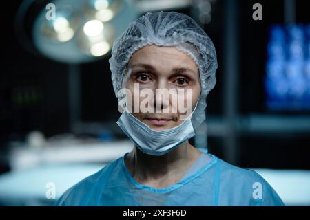 Ritratto in vita di una donna anziana come chirurgo che guarda la macchina fotografica in piedi in sala operatoria e indossa maschera, spazio per le copie Foto Stock