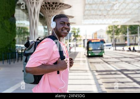 Un giovane africano che indossa cuffie e uno zaino sorride mentre cammina per la strada. Ha uno zaino verde e una camicia rosa Foto Stock