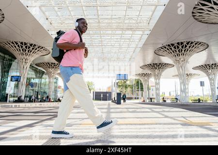 Un uomo che indossa una camicia rosa e un jeans cammina lungo un marciapiede con uno zaino. Sta ascoltando musica con le sue cuffie Foto Stock