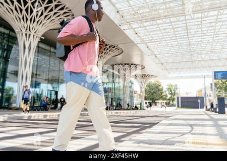 Un uomo con una camicia rosa e un jeans blu cammina lungo un marciapiede con uno zaino. Sta ascoltando musica con le sue cuffie Foto Stock