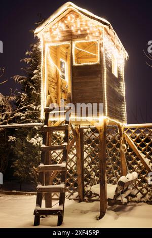 Una casa sull'albero di legno in una notte d'inverno - coperta di neve e illuminata con molte luci natalizie. Foto Stock