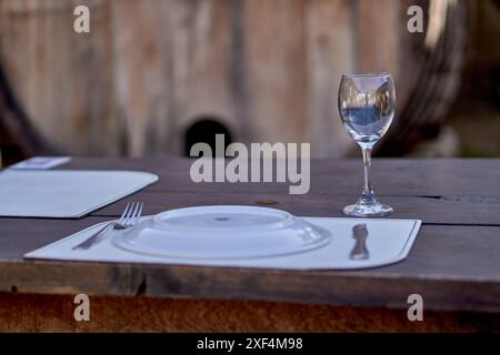 piatto con posate vuote e vetro su un tavolo di legno con un barile sullo sfondo. Pranzo presso il ristorante nel cortile di un'azienda vinicola. Foto Stock