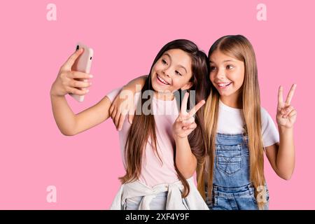 Foto ravvicinata due bambine la sua bionda bruna ragazze lunghe e graziose telefonate per capelli fanno selfie per la mamma mostra la V-sign indossando jeans casual Foto Stock