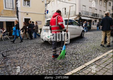 Spazzatrice stradale in uniforme rossa con una scopa verde su una strada acciottolata mentre i pedoni camminano nel quartiere storico di Alfama Foto Stock