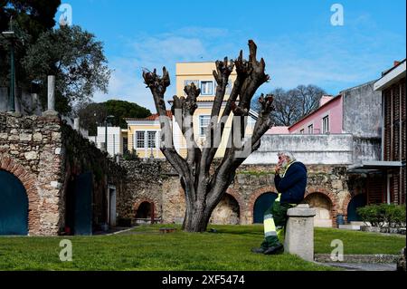 Custode che fuma in uno dei cortili di Castelo de São Jorge, con un albero potato e storiche mura di pietra sullo sfondo Foto Stock