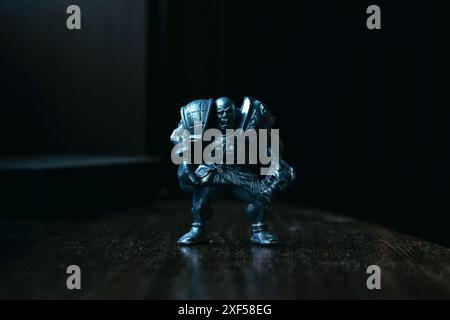 Una statuetta di un guerriero calvo aggressivo e vizioso in armatura con un teschio si erge su un tavolo di legno ed è illuminata da una luce fredda su un dorso scuro Foto Stock