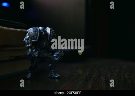 Una statuetta di un guerriero calvo aggressivo e vizioso in armatura con un teschio si erge su un tavolo di legno accanto a libri e luci su uno sfondo scuro Foto Stock