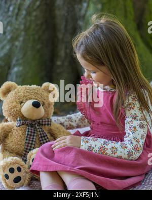 Primo piano ritratto di una bambina carina. Un bambino siede vicino a un grande albero, accanto a un grande giocattolo, orsacchiotto. Ragazza che guarda la sua amica Foto Stock