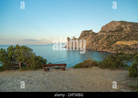 Una panchina è seduta su una spiaggia rocciosa accanto all'oceano. Il cielo è blu e il sole sta tramontando. Foto Stock