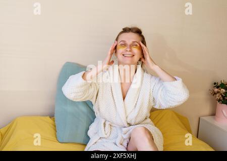 Una donna è seduta su un letto con una coperta gialla e una veste bianca. Indossa una maschera per gli occhi gialla e ha un rivestimento per gli occhi giallo. Sta sorridendo e sh Foto Stock