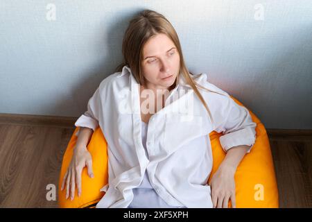 Una donna è seduta su un cuscino giallo sul pavimento. Indossa una camicia bianca e ha la testa abbassata. La scena è triste e solitaria Foto Stock