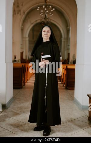 Scatto verticale di una suora caucasica matura che tiene in mano un libro della Bibbia in piedi al nartece nella chiesa cattolica, guardando la macchina fotografica Foto Stock