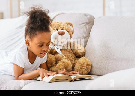 Giovane ragazza che legge Libro con orsacchiotto sul divano Foto Stock