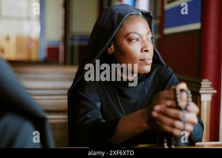 Giovane suora cattolica afroamericana che tiene le perle rosarie sedute sulla pancia e guardando lontano, tiro medio da vicino Foto Stock