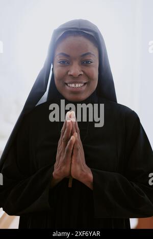 Verticale in vita ritratto di felice suora afroamericana cattolica che posa per la macchina fotografica con le mani in preghiera Foto Stock