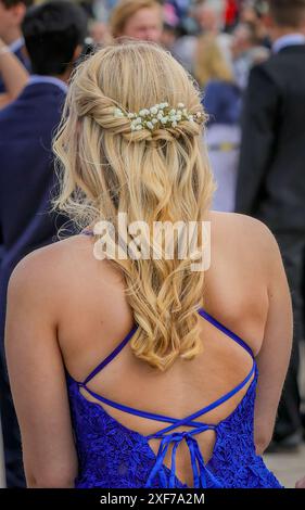 Vista posteriore verticale di una giovane donna con capelli biondi in una acconciatura intrecciata ornata da fiori, che indossa un abito blu in occasione di un ballo all'aperto. Foto Stock