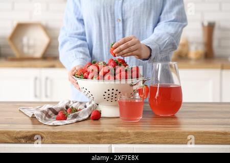 Donna con fragole fresche nel colino e succo di fragole su un tavolo di legno in cucina Foto Stock