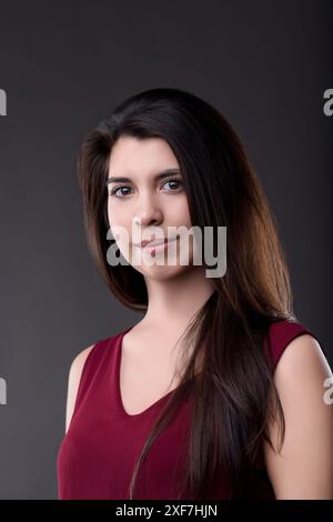 Una giovane donna sicura e felice con lunghi capelli scuri si posa elegantemente in un top Bordeaux su un sereno sfondo grigio in un ambiente da studio Foto Stock