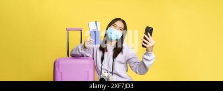 Felice donna coreana che prende selfie con passaporto e biglietti di volo, in vacanza, in piedi vicino alla grande carina valigia rosa, sfondo giallo Foto Stock
